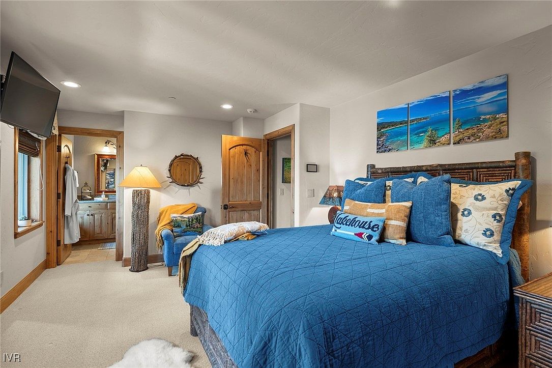 This is a cozy primary bedroom featuring a large bed with a blue quilted cover and decorative pillows, including one that says "Lakehouse". The room has a rustic yet comfortable feel, with wooden accents and a triptych of a lake scene hanging above the bed. The room also features a view into the primary bathroom.