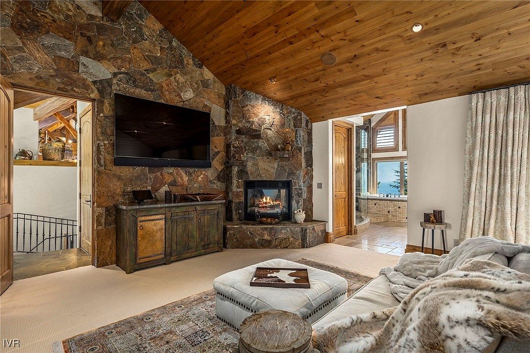 This living room features a stone fireplace with a mounted television above a wooden cabinet. The room has a rustic yet luxurious feel, with a wooden ceiling, a comfortable sofa with a fur throw, and a patterned rug. An open doorway leads to another room with stairs, adding depth and interest to the space.