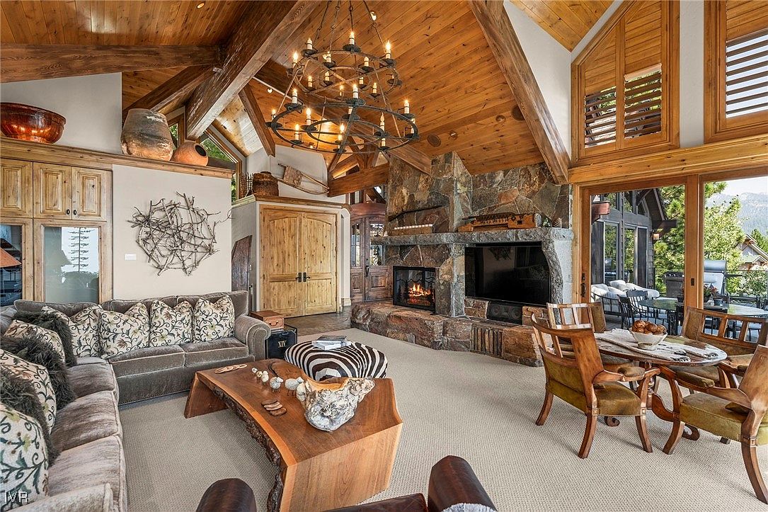 This is an interior shot of a luxurious living room featuring a stone fireplace with a mounted television, a wooden coffee table, and comfortable sofas. The room has high, exposed wooden beam ceilings and a large chandelier, creating a warm and inviting atmosphere. Sliding glass doors lead to an outdoor patio area, enhancing the sense of space and connection to nature.