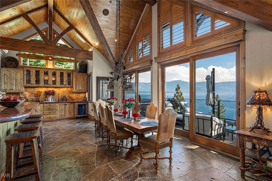 This interior shot showcases a dining area with a rustic yet elegant design. A large wooden dining table is set with flowers, surrounded by upholstered chairs. The room features high ceilings with exposed wooden beams, a unique chandelier, and large windows offering a stunning view of the lake and mountains, creating a luxurious and inviting atmosphere.