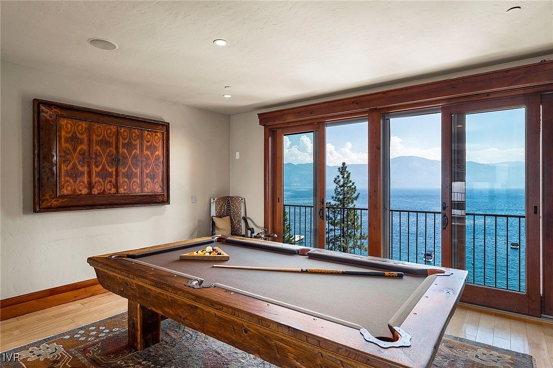 This interior shot showcases a living room featuring a wooden pool table as the focal point, complemented by a large framed artwork on the wall. Expansive sliding glass doors offer a stunning view of a lake and mountains, enhancing the room's appeal. The space is well-lit and exudes a sense of luxury and relaxation.