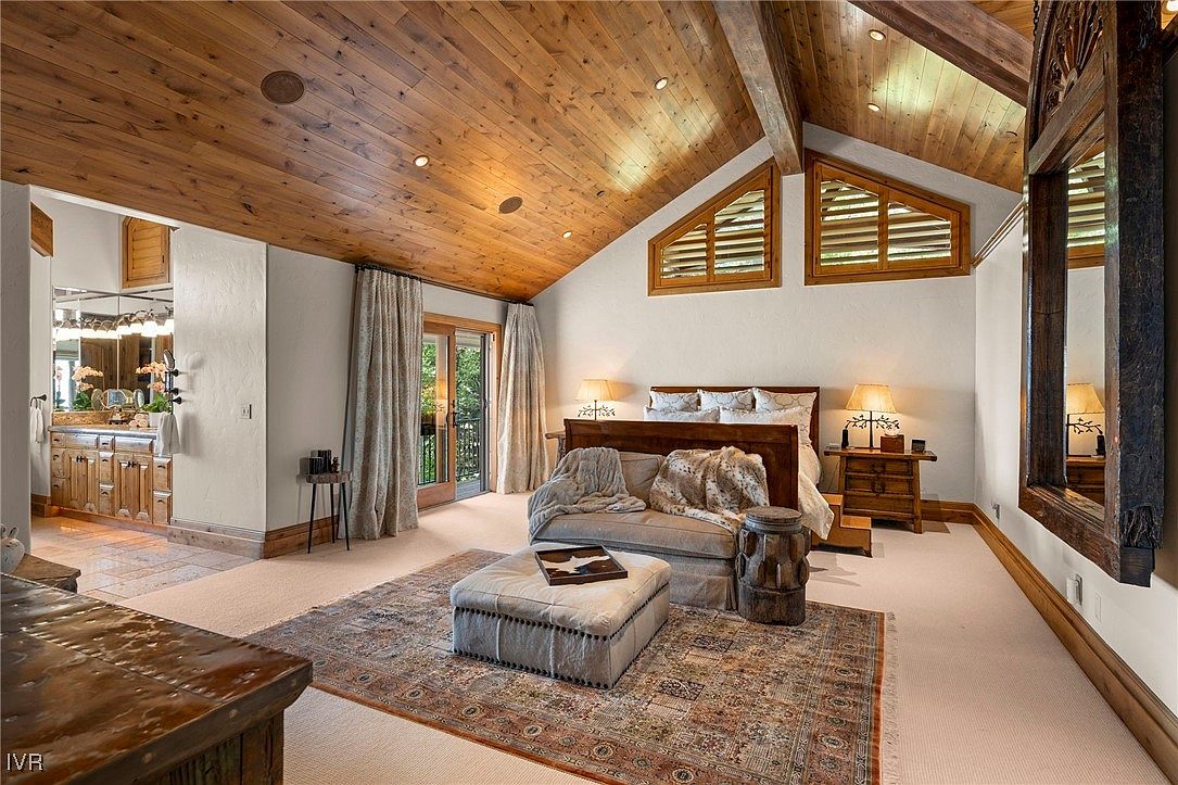 This is a primary bedroom featuring a wooden ceiling, a large bed with white linens, and a seating area with a patterned rug. The room has a rustic yet elegant feel, with natural light coming through the windows and a door leading to a balcony. The overall impression is one of comfort and luxury.