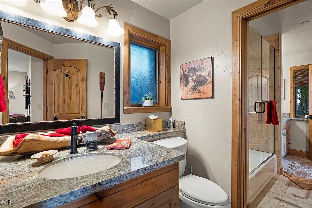 This is a well-appointed bathroom featuring a granite countertop vanity with a wooden base, a large mirror, and a window providing natural light. The bathroom includes a toilet and a glass-enclosed shower. The overall design has a rustic, yet elegant feel, suitable for a guest bathroom in a high-end property.
