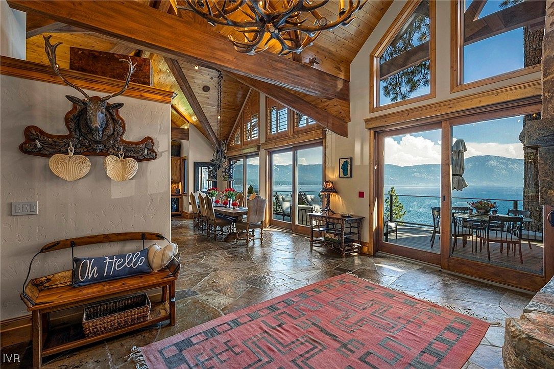 This interior shot showcases a luxurious living room with a rustic yet elegant design. The room features exposed wooden beams, a stone fireplace, and large windows offering stunning lake views. A dining area is visible in the background, and the overall impression is one of spaciousness and high-end comfort.
