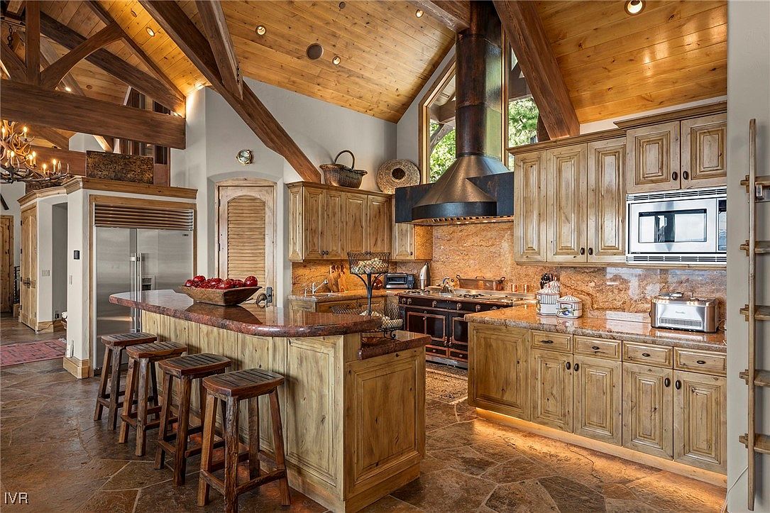 This is a warm and inviting kitchen with rustic charm, featuring wooden cabinetry, granite countertops, and a large island with bar seating. The high, beamed ceilings and stone flooring add to the home's character, while modern appliances provide convenience. The kitchen is well-lit and appears spacious, perfect for cooking and entertaining.