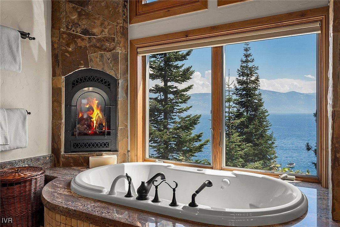 This is a luxurious primary bathroom featuring a large soaking tub positioned in front of a window with a scenic view of a lake and trees. A stone fireplace adds warmth and ambiance to the space. The bathroom is designed with high-end fixtures and finishes, creating a spa-like atmosphere.