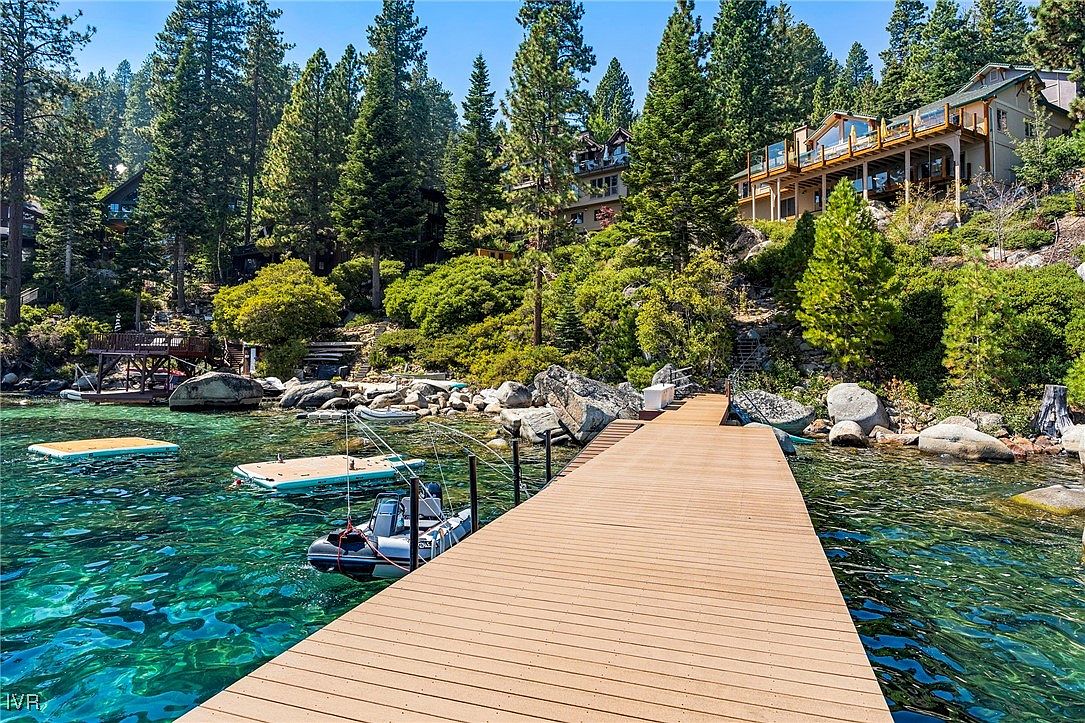 This image showcases a stunning waterfront property with a long wooden dock extending into crystal-clear turquoise water. A boat is moored alongside the dock, and in the background, luxurious homes are nestled among lush green trees on a hillside. The scene evokes a sense of tranquility and upscale lakeside living.