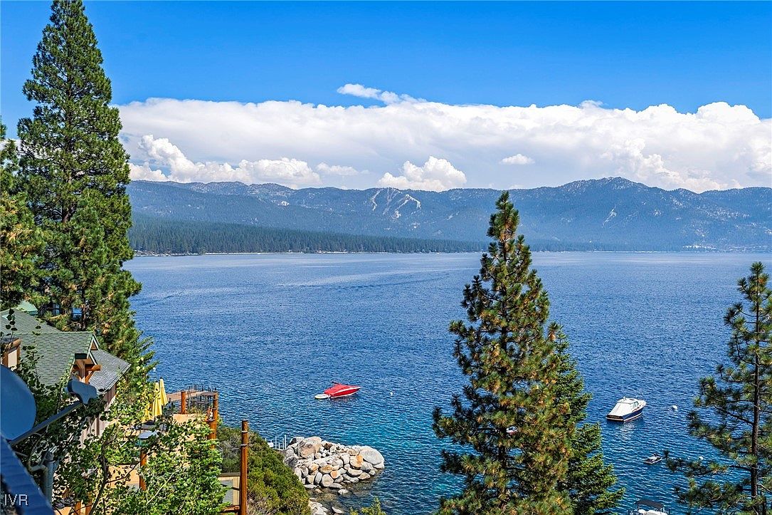 This image showcases a stunning lake view from a patio or deck, framed by tall pine trees. The clear blue water is dotted with boats, and mountains rise in the background under a partly cloudy sky. The scene evokes a sense of tranquility and natural beauty, highlighting the property's prime location and outdoor living potential.