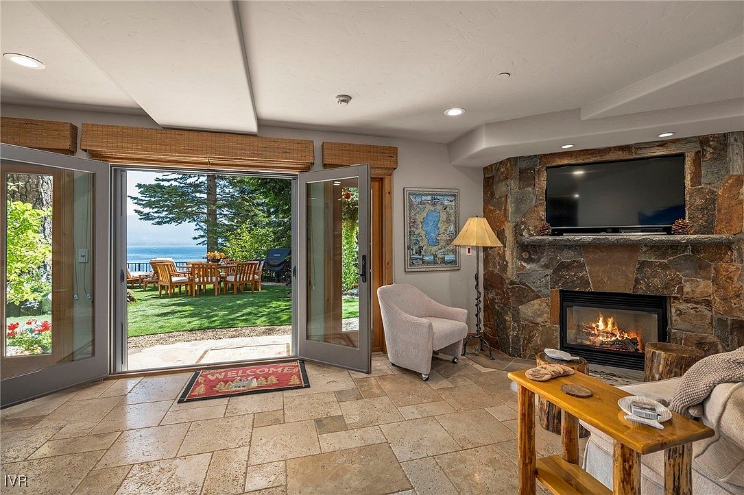 This inviting living room features a stone fireplace with a mounted television, creating a cozy focal point. Large glass doors open to an outdoor patio with a view of the lake, seamlessly blending indoor and outdoor living. The room is furnished with comfortable seating and a rustic wooden side table, enhancing the warm and welcoming atmosphere.