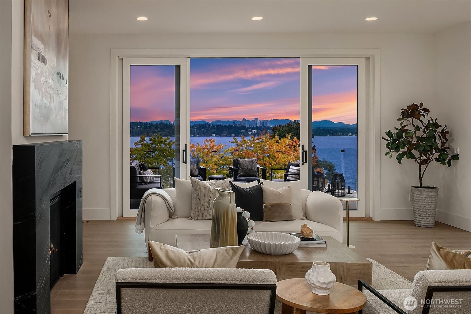 This is a beautifully staged living room with a focus on the stunning view through large sliding glass doors. The neutral color palette, comfortable seating arrangement, and modern fireplace create an inviting and luxurious atmosphere. The room is well-lit and appears spacious, emphasizing the connection between indoor and outdoor living.