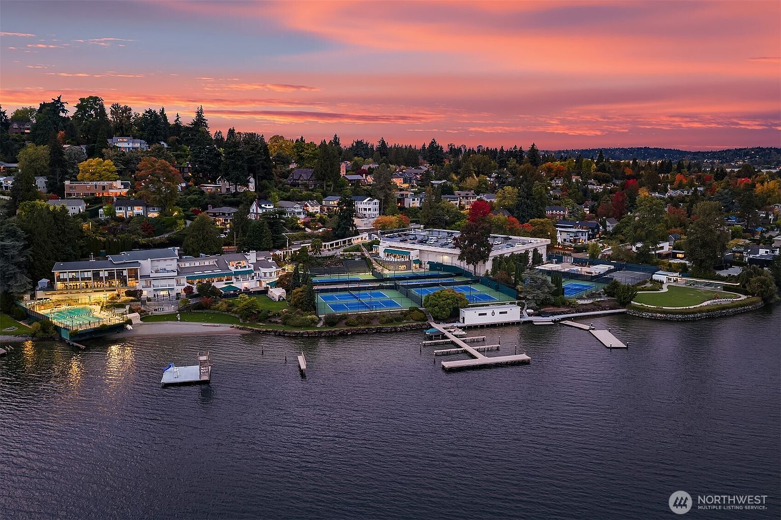 This aerial shot showcases a luxurious waterfront property at sunset, featuring multiple tennis courts, a private beach, and a large building complex with a pool. The surrounding neighborhood is lush with mature trees, and the overall impression is one of exclusivity and high-end living. The water is calm and reflects the colors of the sky.