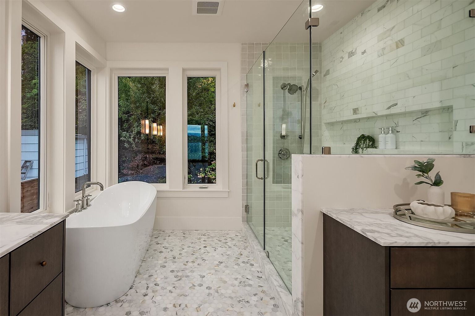 This is a bright and modern primary bathroom featuring a freestanding white bathtub positioned near large windows that offer a view of the outdoors. The bathroom includes a glass-enclosed shower with marble-like tiling and a built-in shelf. The floor is covered in a decorative mosaic tile pattern, and the vanity has a dark wood finish with a marble countertop, creating a luxurious and spa-like atmosphere.