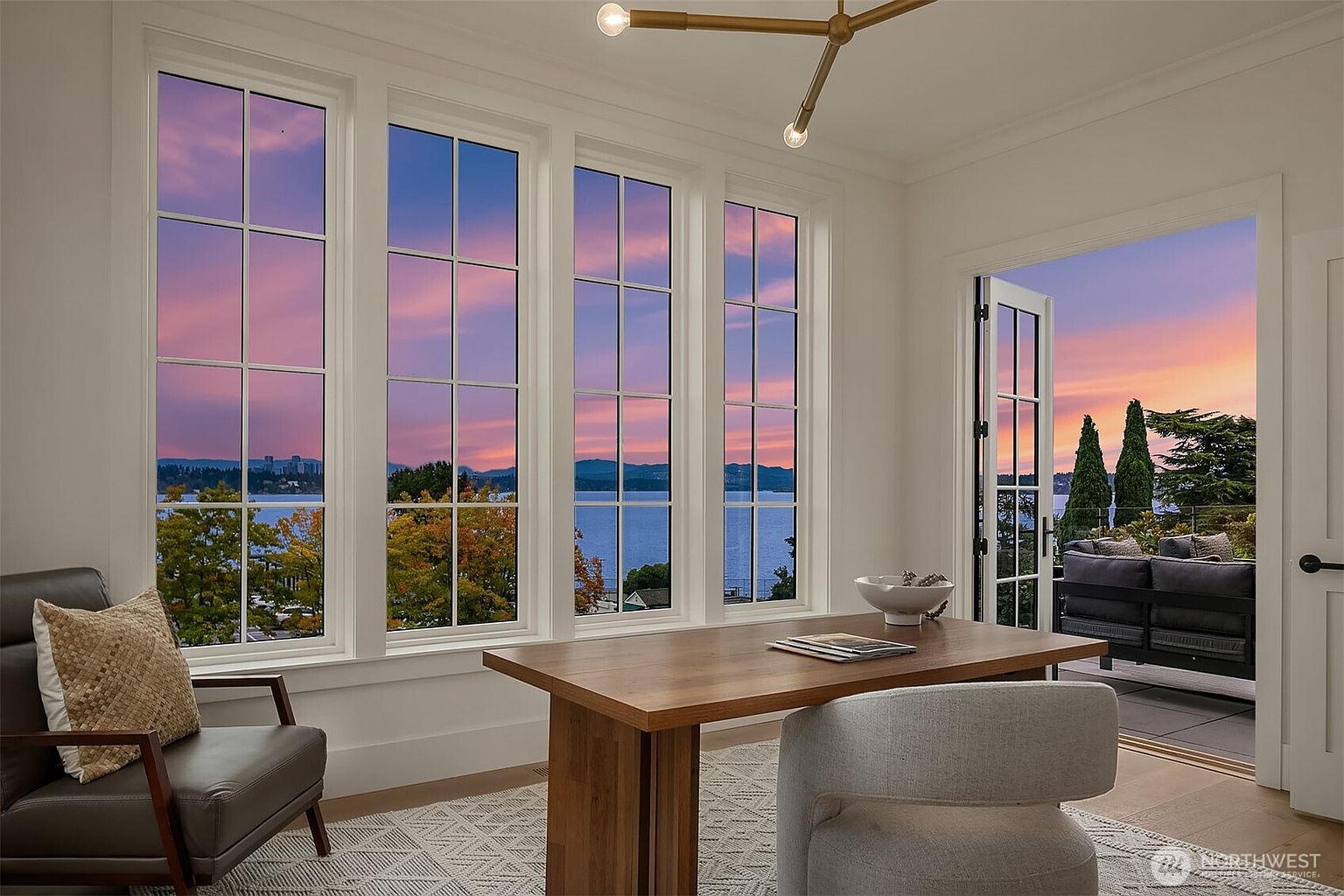 This is an interior shot of a bright and airy office space. The room features a large wooden desk, a comfortable chair, and a wall of windows offering a stunning view of a lake at sunset. An open door leads to an outdoor patio with comfortable seating, creating a seamless indoor-outdoor living experience.