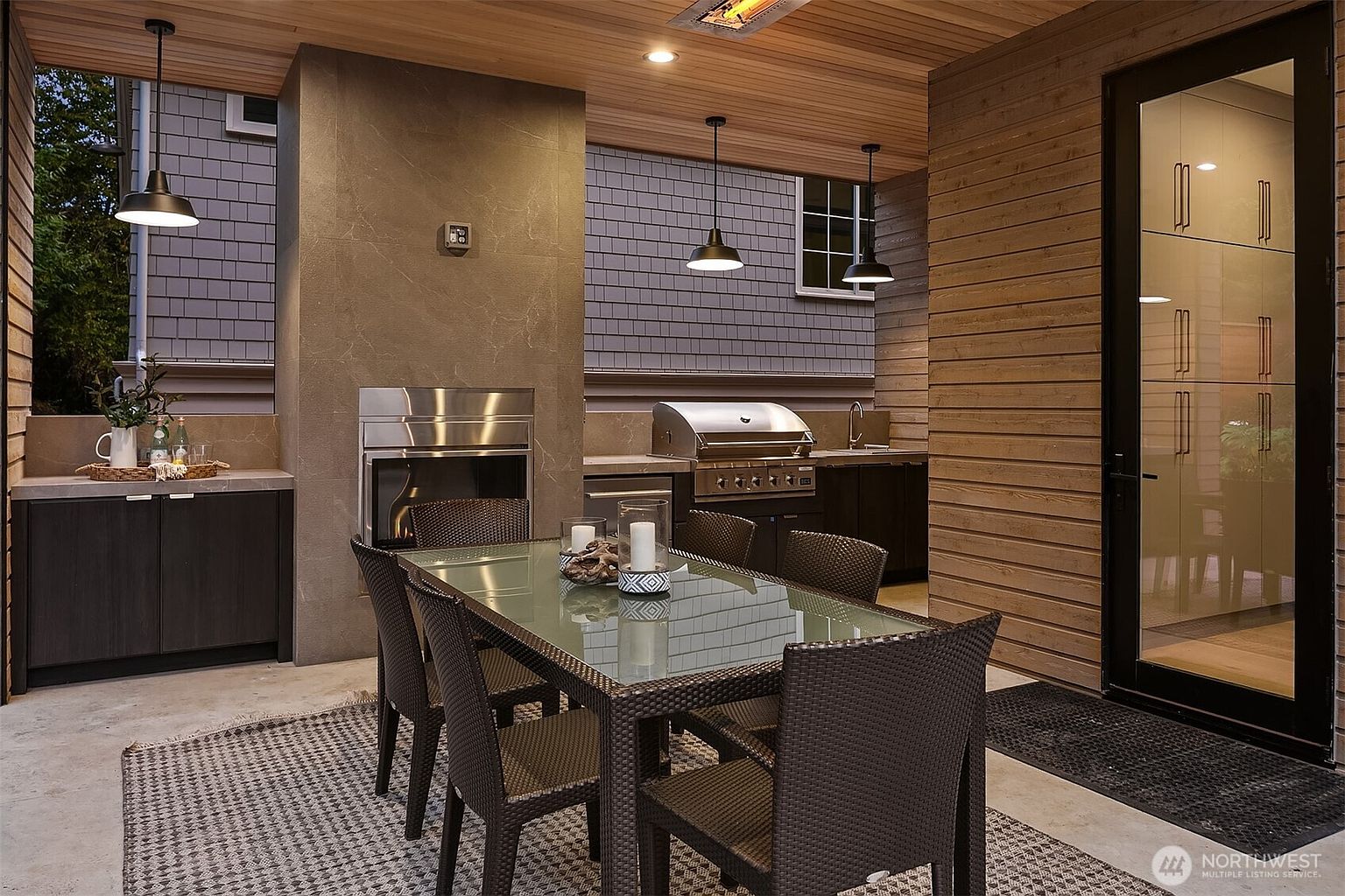 This image showcases a well-appointed outdoor patio area, featuring a dining table with seating for six, a built-in grill and fireplace, and stylish cabinetry. The space is covered with a wooden ceiling and features modern lighting fixtures, creating an inviting atmosphere for outdoor dining and entertaining. The patio is furnished with a patterned rug and is accessible through a glass door.