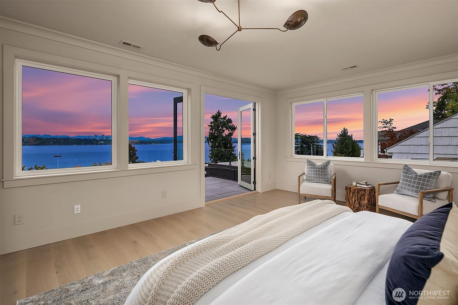 This is a bright and airy primary bedroom featuring large windows with stunning sunset views over the water. The room is decorated in a neutral palette with light wood floors, white walls, and comfortable seating. A plush bed with a white duvet and blue accent pillows completes the serene and inviting atmosphere.