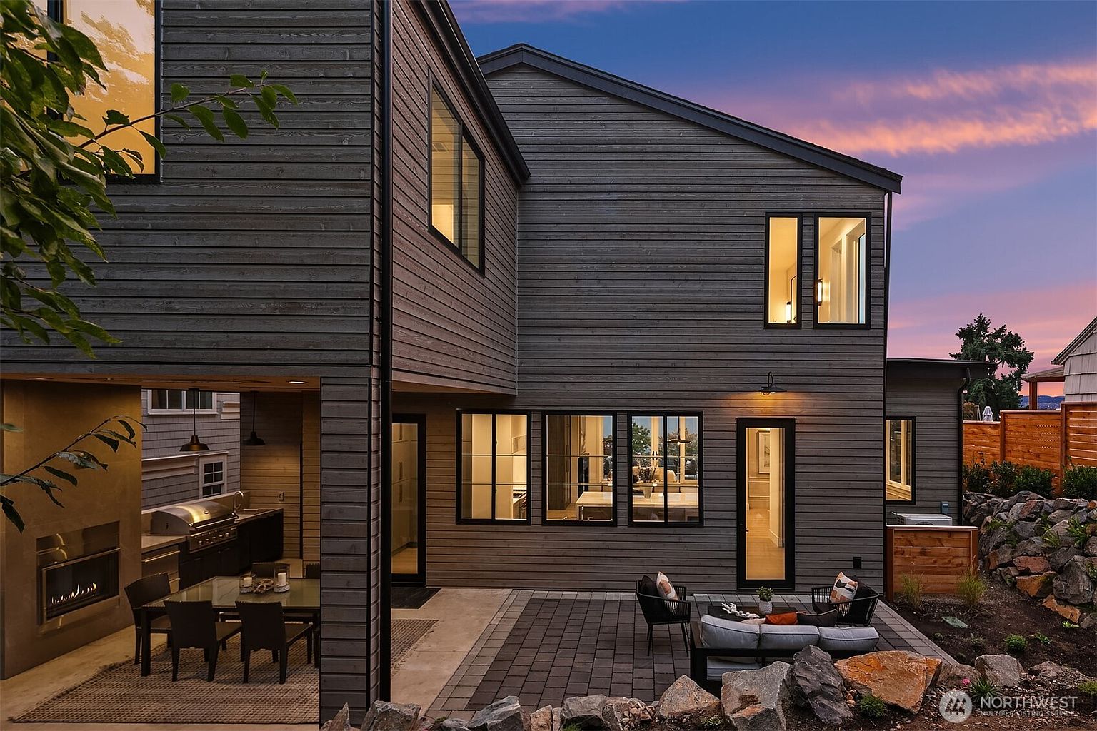 This is a rear exterior view of a modern home, showcasing a dark horizontal wood siding and black-framed windows. The outdoor space features a patio with comfortable seating and a built-in outdoor kitchen area with a grill and fireplace. The landscaping includes rock features and plantings, creating an inviting and stylish backyard.