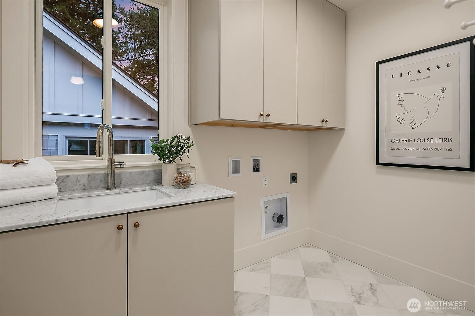 This is a well-lit laundry room featuring a neutral color palette with white and beige tones. The room includes upper cabinets, a sink with a marble countertop, and a washer hookup. The flooring is a light-colored tile with a diamond pattern, and a framed art print adds a touch of sophistication to the space.