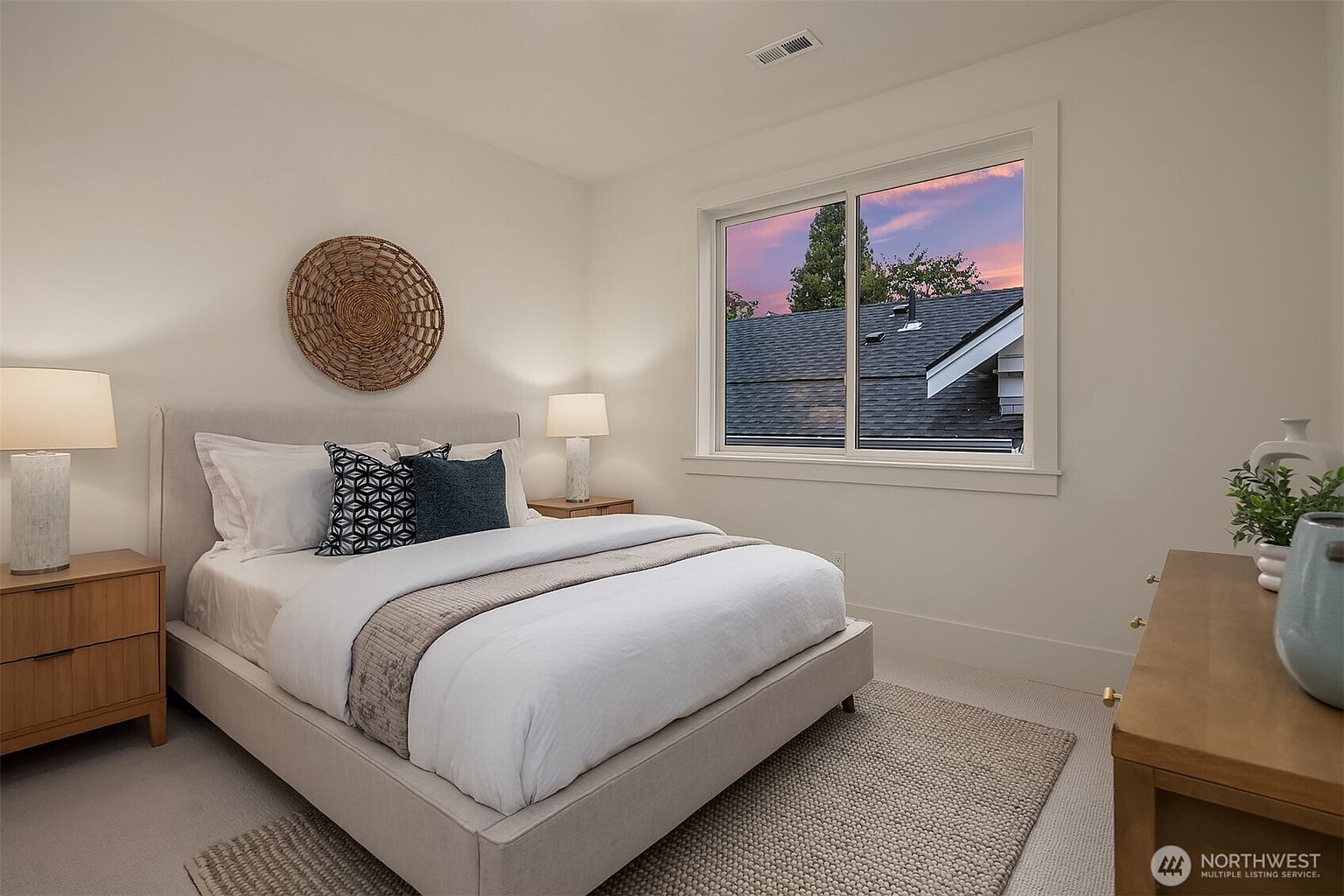This is a cozy bedroom featuring a neutral color palette with a bed dressed in white linens and decorative pillows. A woven basket hangs above the bed, adding a touch of texture. The room is well-lit by natural light from a window, creating a serene and inviting atmosphere.