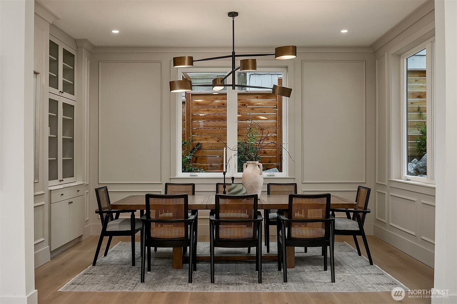 This is an interior shot of a dining room featuring a long wooden table with black and cane chairs. A modern chandelier hangs above the table, and a large window provides natural light. The room is decorated in neutral tones with paneled walls, creating a sophisticated and inviting atmosphere.