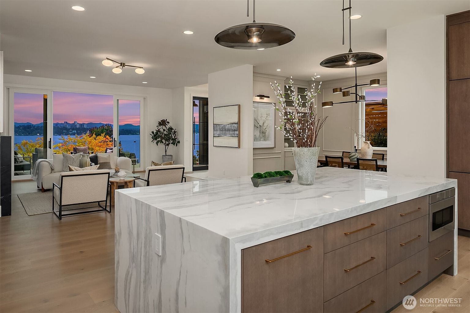 This is a bright and modern kitchen with a large marble island as the focal point. The island features wooden cabinets with gold hardware and a built-in microwave. The kitchen seamlessly flows into the living and dining areas, offering stunning views of the lake and surrounding landscape through large windows, creating an open and inviting atmosphere.