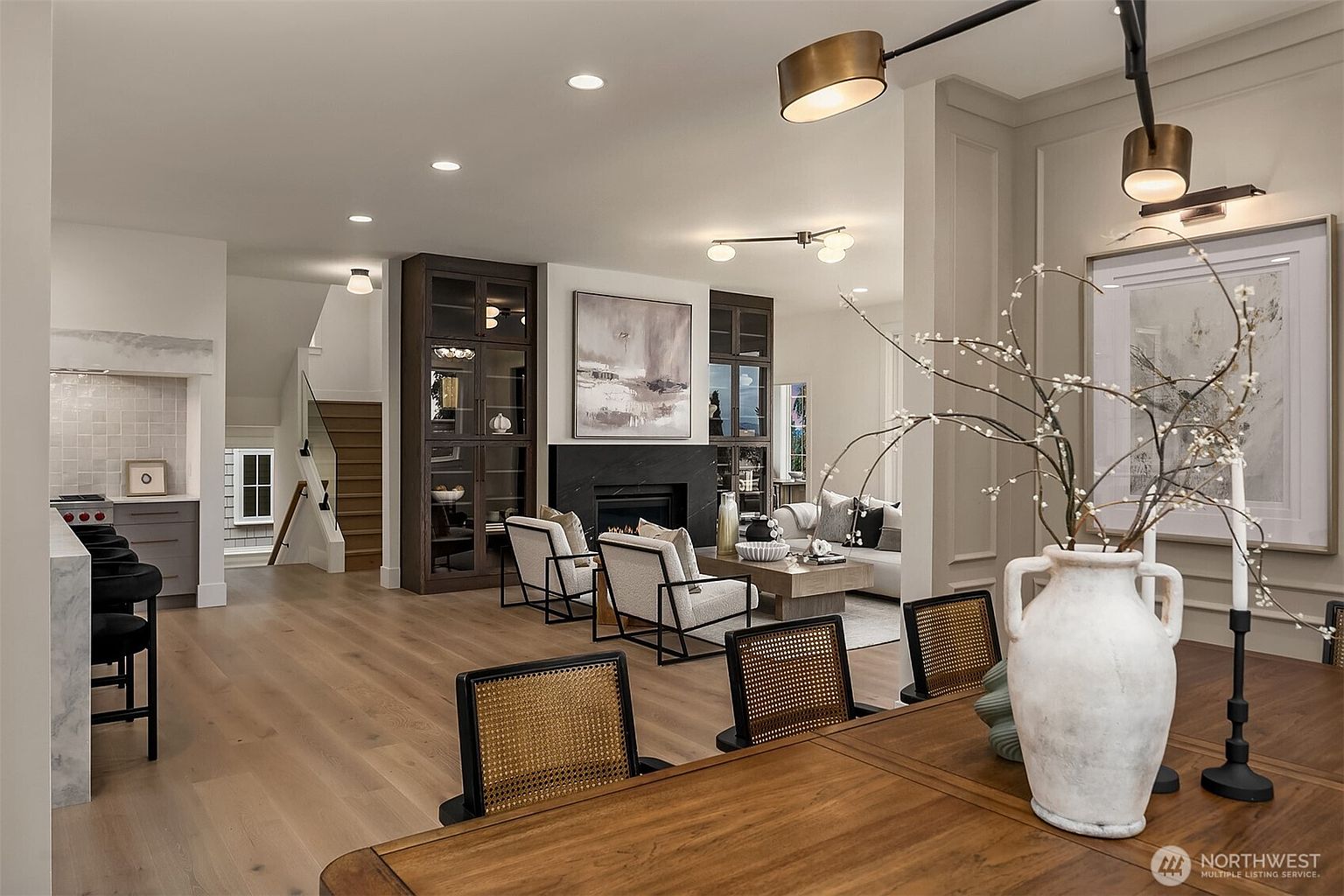 This interior shot showcases a stylish living room with hardwood floors, a fireplace flanked by built-in cabinets, and modern furniture. The open-concept design flows into the dining area, highlighted by a wooden table and unique chairs. The room is well-lit with a combination of recessed lighting and decorative fixtures, creating a warm and inviting atmosphere.