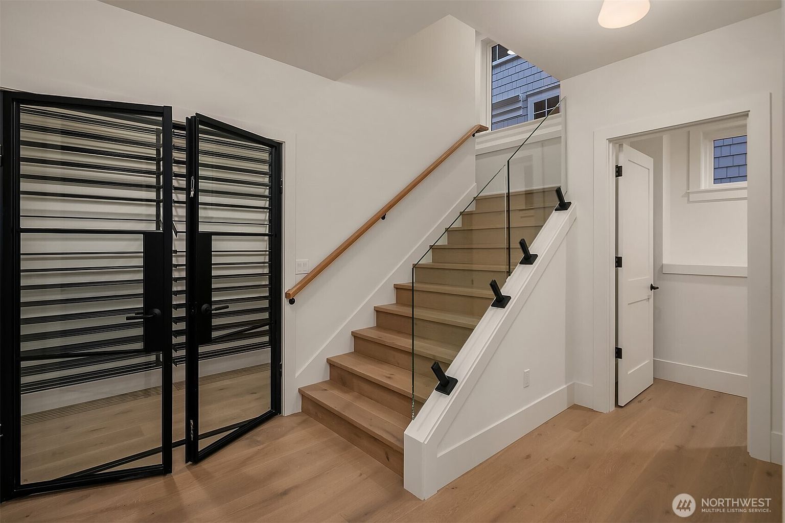 This interior shot showcases a modern hallway with wooden stairs and a glass railing. A striking black metal gate adds a unique architectural element. The space is well-lit and features light wood flooring, contributing to a clean and contemporary aesthetic.