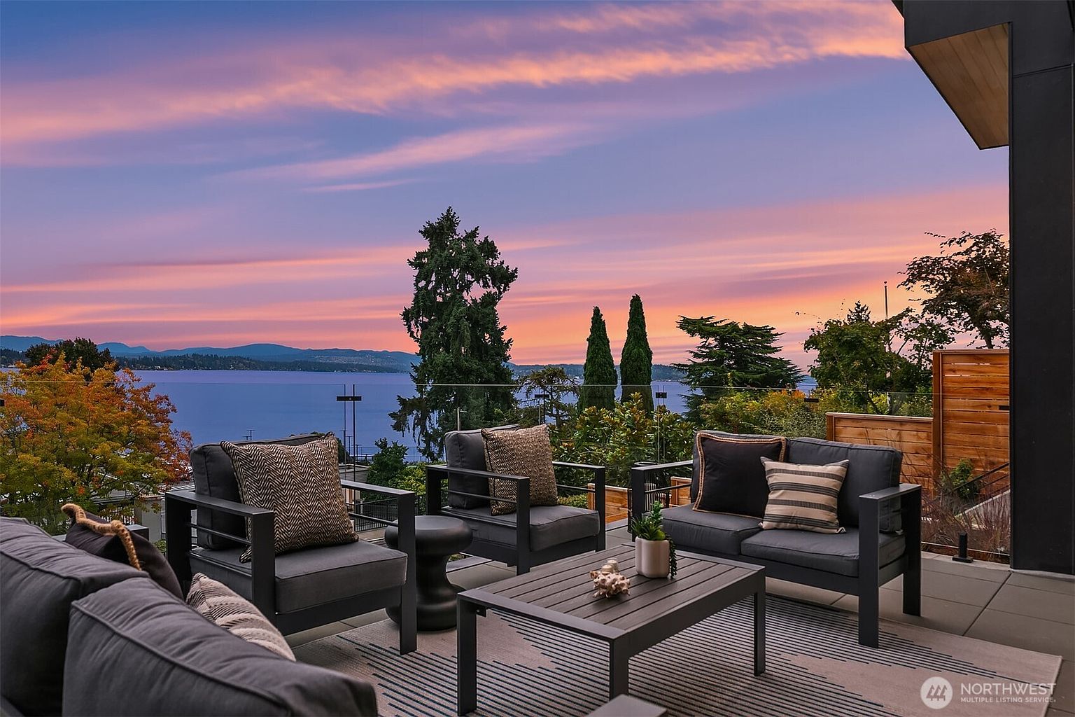 This image showcases a luxurious outdoor patio or deck area, furnished with modern, dark-framed seating arrangements and a coffee table, all set upon a striped outdoor rug. The space offers a stunning panoramic view of a lake or ocean at sunset, with a vibrant sky painted in hues of pink and orange, creating an inviting and serene atmosphere perfect for relaxation and entertainment.