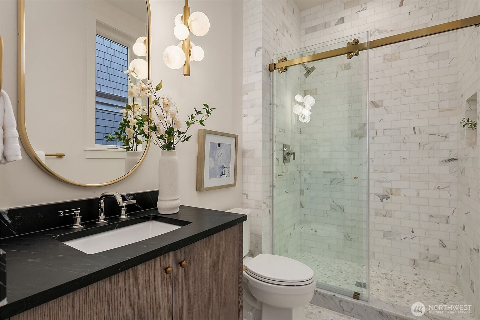 This is a well-lit bathroom featuring a modern design with a vanity, toilet, and glass-enclosed shower. The vanity has a dark countertop and wood-grain cabinets, complemented by a gold-framed mirror and a decorative light fixture. The shower is tiled with white marble-like tiles and has a gold sliding door, creating a luxurious and clean aesthetic.