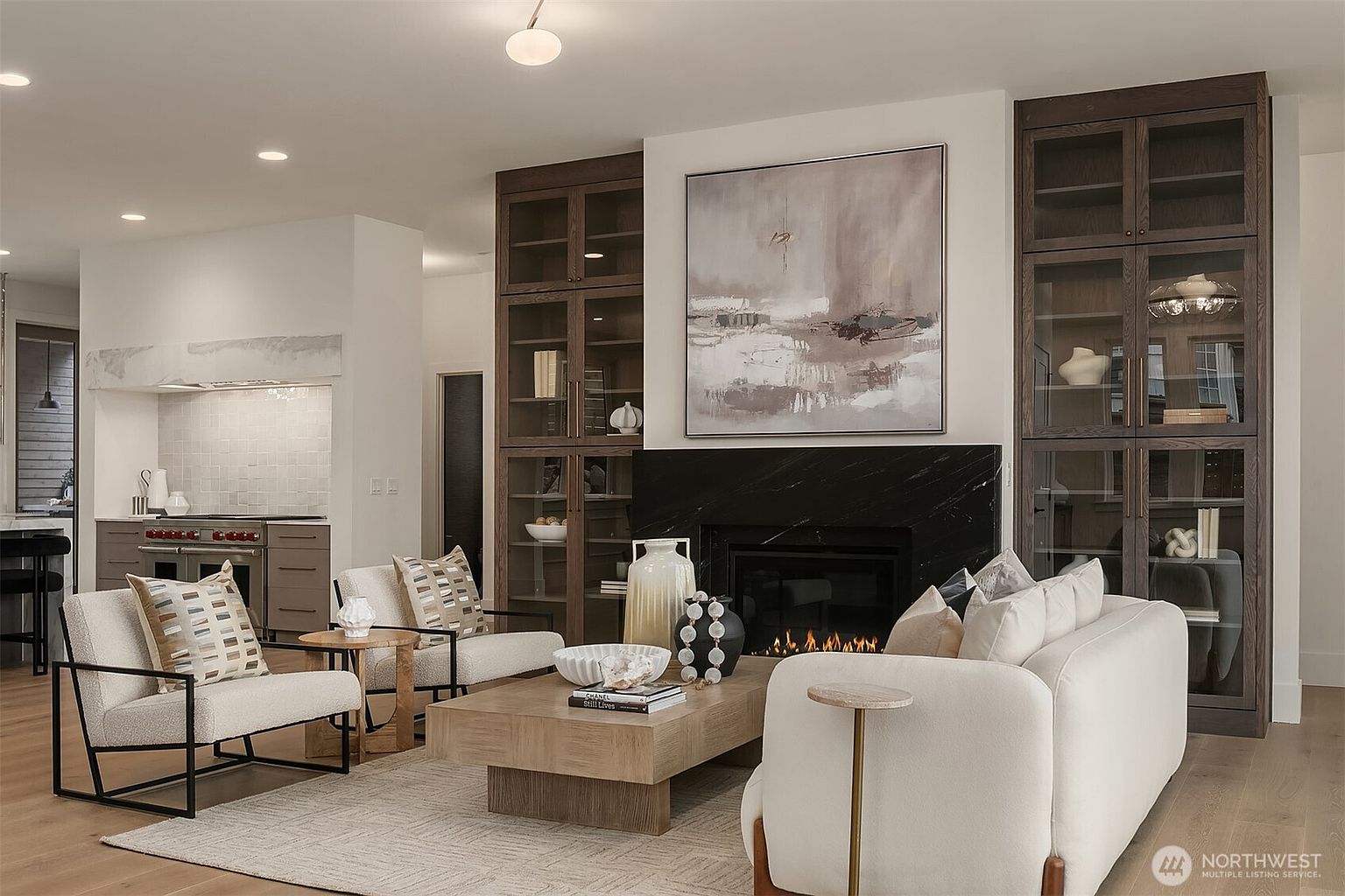 This is an interior shot of a living room featuring a fireplace with a black marble surround, flanked by built-in cabinets with glass doors. The room is furnished with a light-colored sofa and armchairs, arranged around a wooden coffee table. The style is modern and sophisticated, with a neutral color palette and natural light.