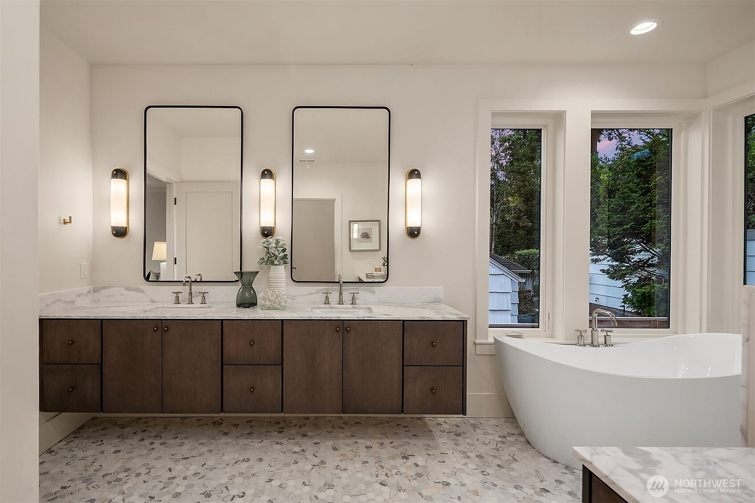 This is a bright and modern primary bathroom featuring a double vanity with dark wood cabinets and marble countertops. Two rectangular mirrors are flanked by vertical sconce lighting. A freestanding white bathtub sits beneath a window offering natural light and a view of the outdoors, and the floor is covered in a stylish mosaic tile pattern.