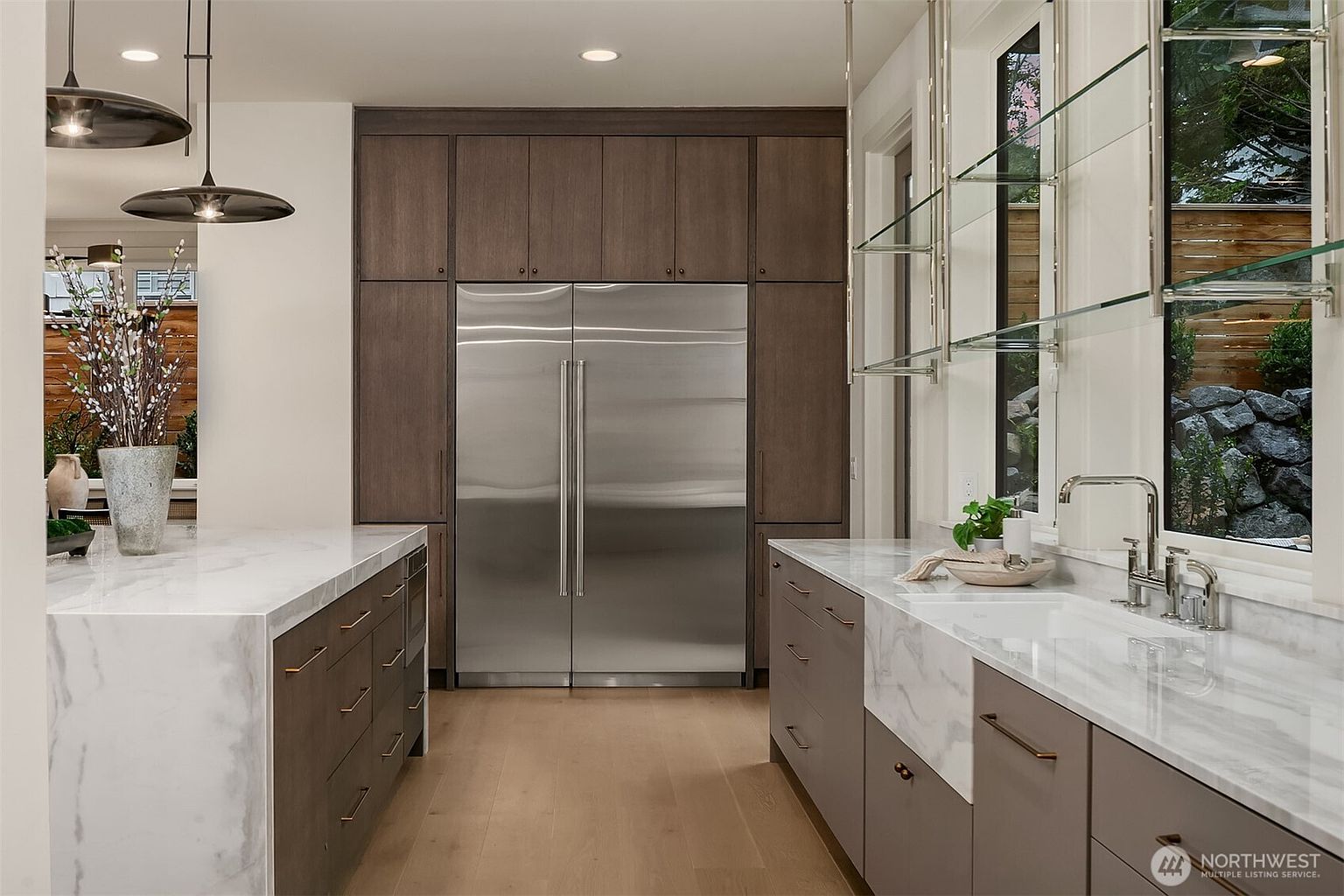 This is a well-lit kitchen featuring a large stainless steel refrigerator integrated into dark wood cabinetry. The kitchen also has marble countertops on the island and sink area, with modern fixtures and hardware. The overall impression is one of contemporary elegance and functionality.