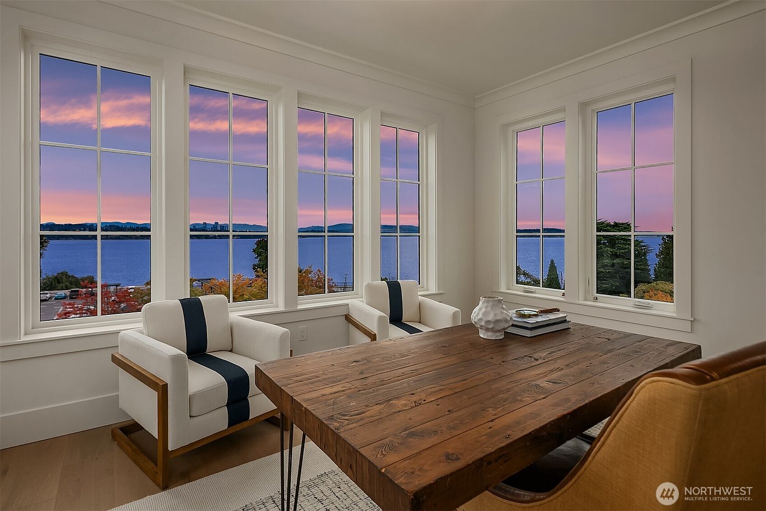 This interior shot showcases a bright and airy office space with a stunning water view through large windows. The room features two armchairs with a unique stripe design, a large wooden desk with books and a decorative vase, and hardwood floors. The overall impression is one of sophistication and tranquility, ideal for a productive workspace.