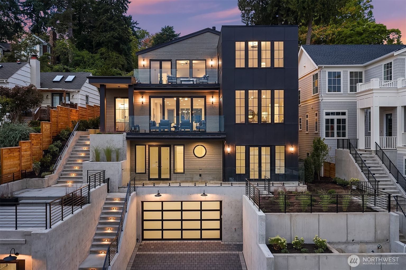 This is a striking front view of a modern, multi-level home featuring a unique architectural design. The facade combines wood siding with a dark, contemporary tower element, complemented by glass-railed balconies. The property includes a garage accessed via a driveway and multiple sets of stairs leading to the upper levels, enhanced by subtle lighting.