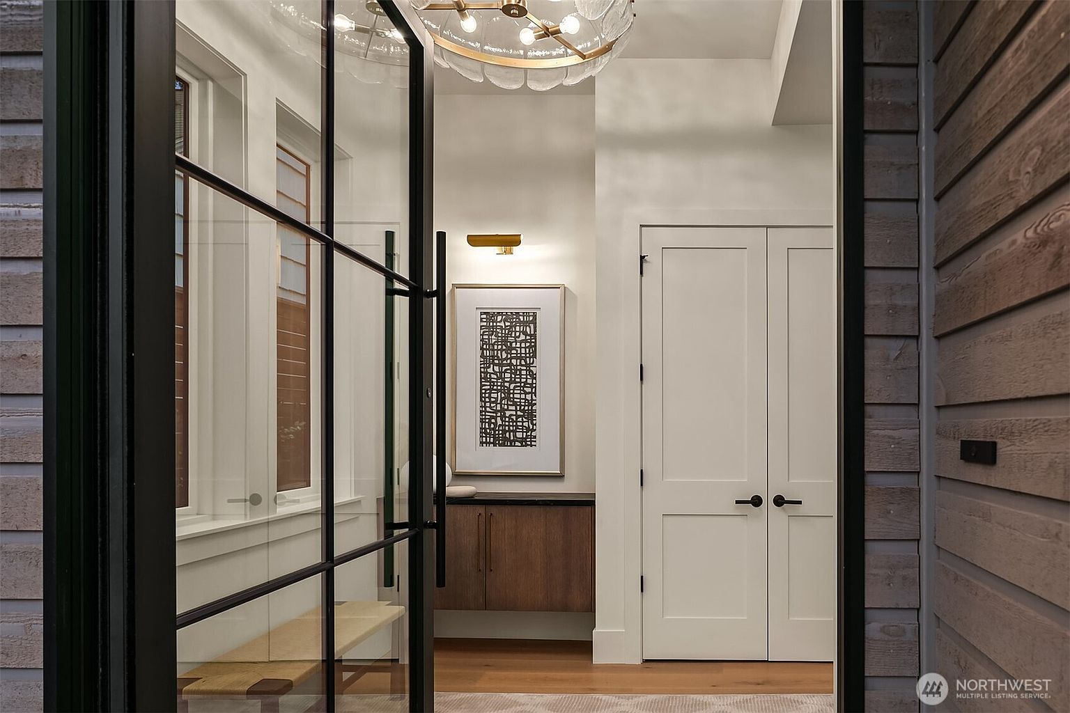 The image showcases an elegant entryway featuring a modern glass and black metal door, leading into a hallway. Inside, there's a stylish light fixture, a framed piece of art above a wooden cabinet, and a set of white double doors. The flooring appears to be hardwood with a subtle rug, creating a welcoming and sophisticated ambiance.