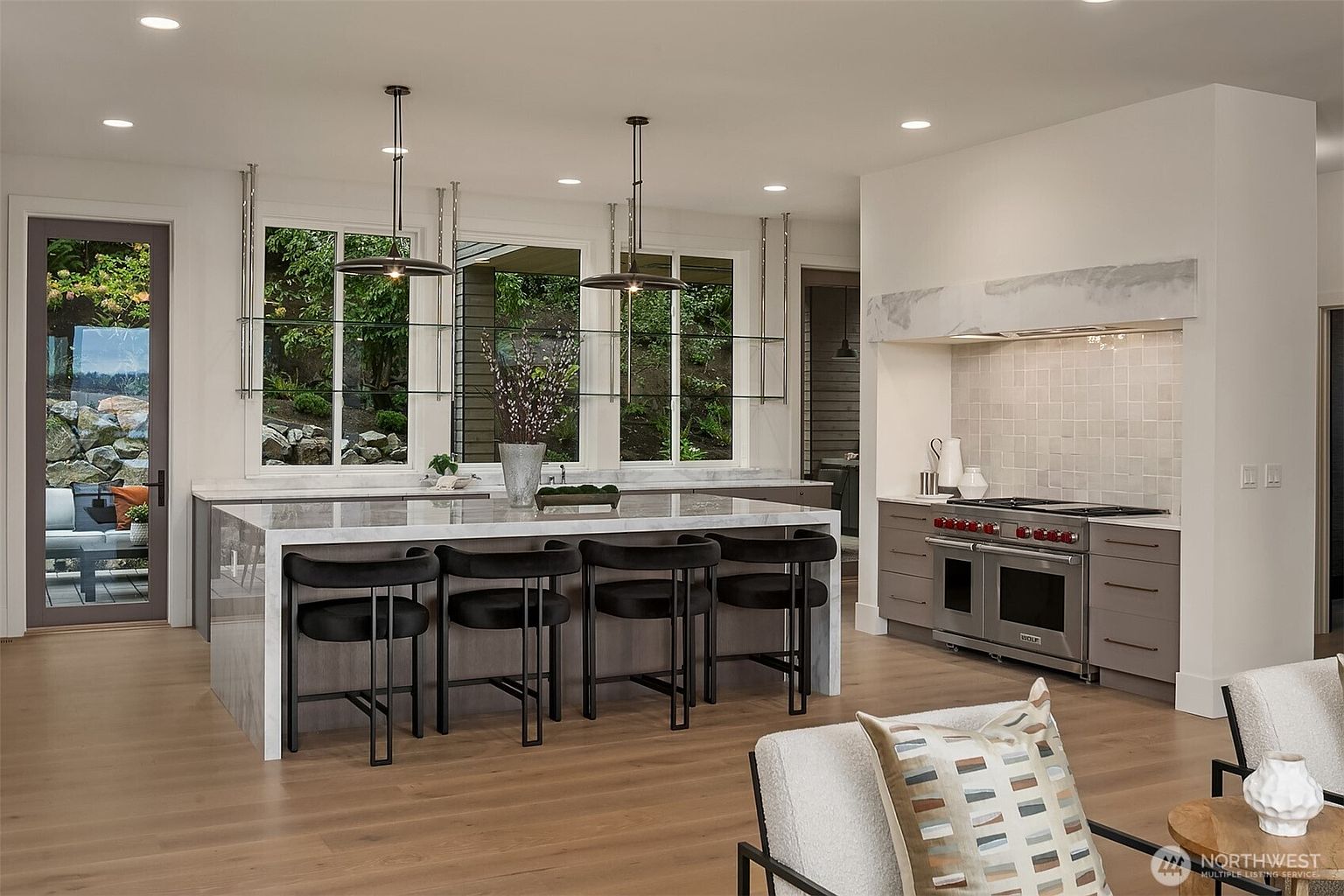 This is a bright and modern kitchen featuring a large island with seating, pendant lighting, and a high-end range with a custom hood. The kitchen has a clean, contemporary design with a neutral color palette and natural wood flooring. Large windows provide ample natural light and views of the outdoor greenery.