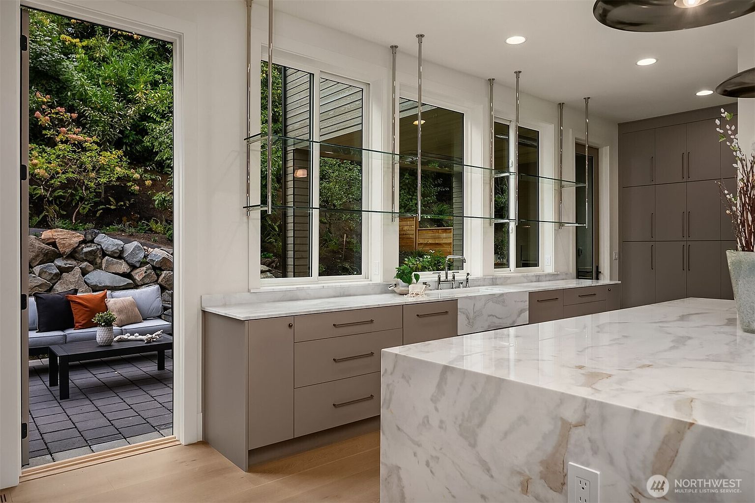 This is a bright and modern kitchen with sleek, taupe-colored cabinetry and marble countertops. Large windows provide ample natural light and a view of the outdoor patio area. The kitchen features stainless steel accents and a spacious island, creating an inviting and functional space.