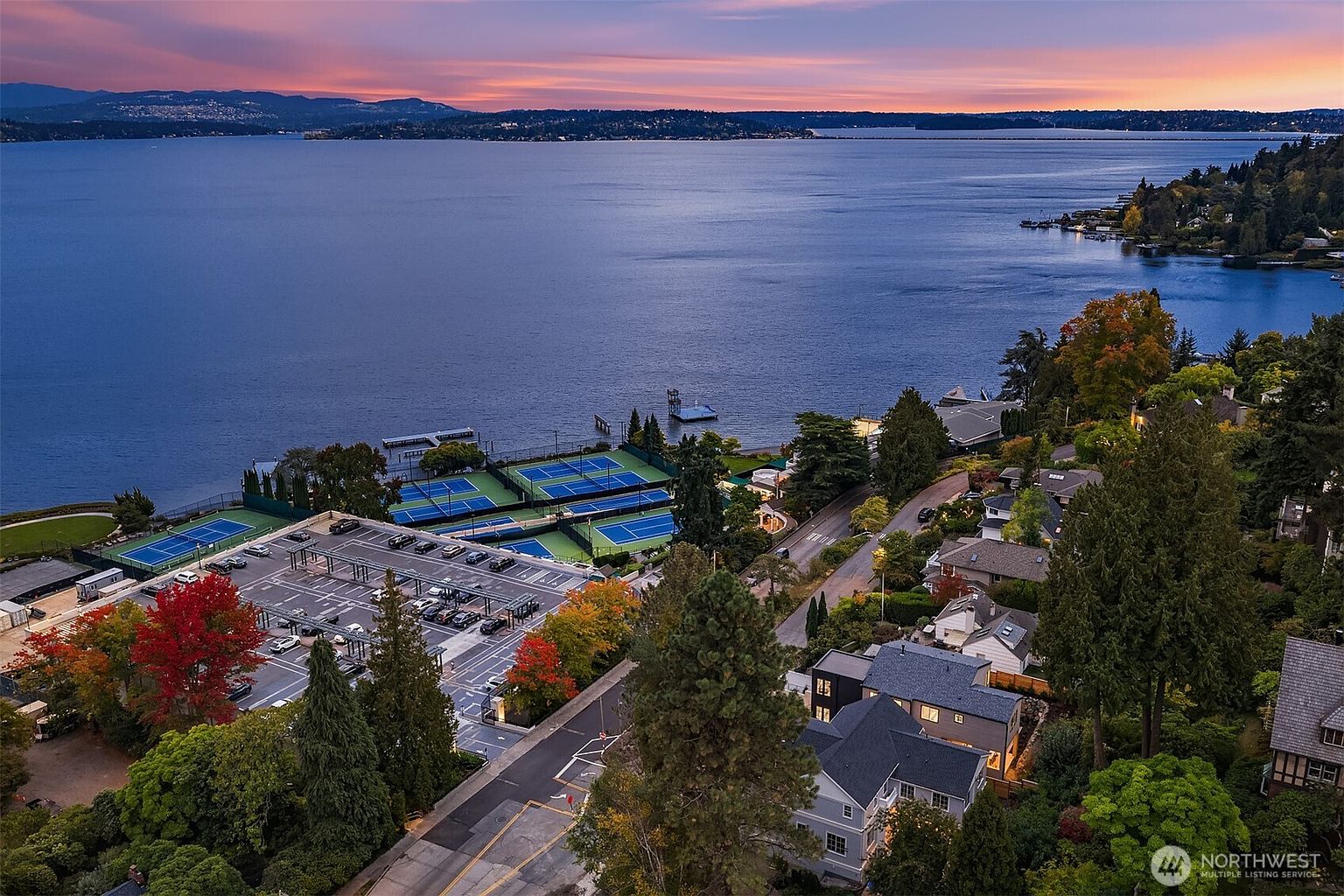 This aerial shot showcases a stunning property with a view of a large body of water at sunset. The property features tennis courts, a parking lot, and well-maintained homes surrounded by lush greenery with trees displaying fall colors. The overall impression is one of luxury and tranquility.