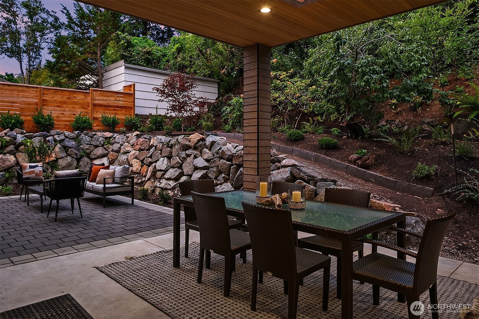 This image showcases a beautifully designed outdoor patio area, perfect for entertaining or relaxing. The space features a dining table with chairs, a lounge area with comfortable seating, and a well-maintained garden backdrop with a stone retaining wall. The patio is partially covered, providing shade and protection from the elements, enhancing its appeal as an outdoor living space.