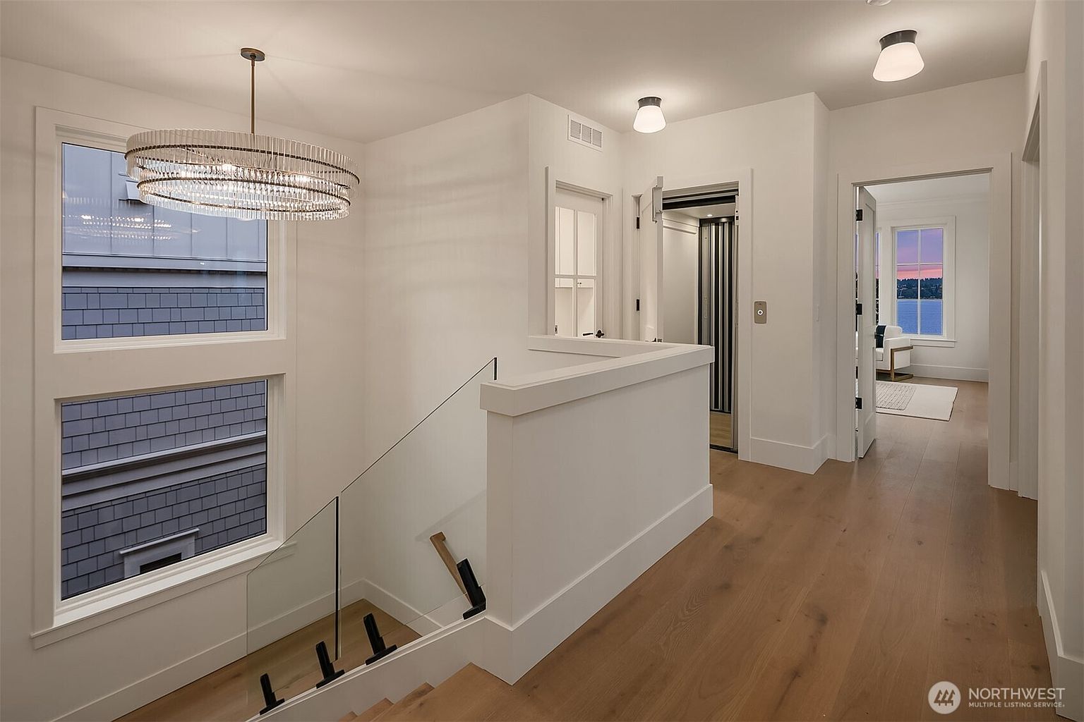 This interior shot showcases a bright and modern hallway with hardwood flooring and white walls. A staircase with glass railings is visible, along with a stylish chandelier. An elevator is present, and a glimpse into a room with a view of the water can be seen through an open doorway, creating an inviting and luxurious atmosphere.