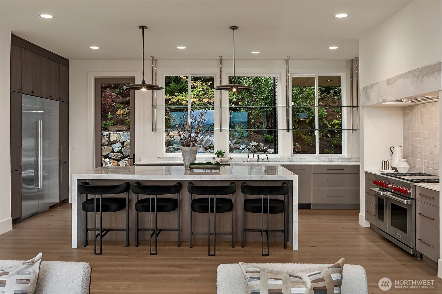This is a bright and modern kitchen featuring a large island with seating, stainless steel appliances, and sleek cabinetry. The kitchen is well-lit with pendant lights and recessed lighting, and large windows offer a view of the lush greenery outside. The overall impression is one of luxury and functionality.