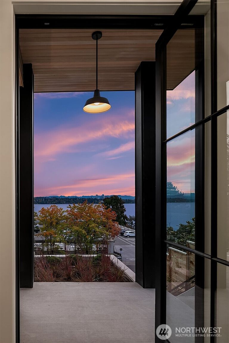 This image showcases a stylish patio or balcony with a stunning view of a lake and a colorful sunset. The space is framed by modern black window frames and features a contemporary light fixture hanging from the wooden ceiling. The view includes trees, a road, and the lake, creating a serene and inviting atmosphere.