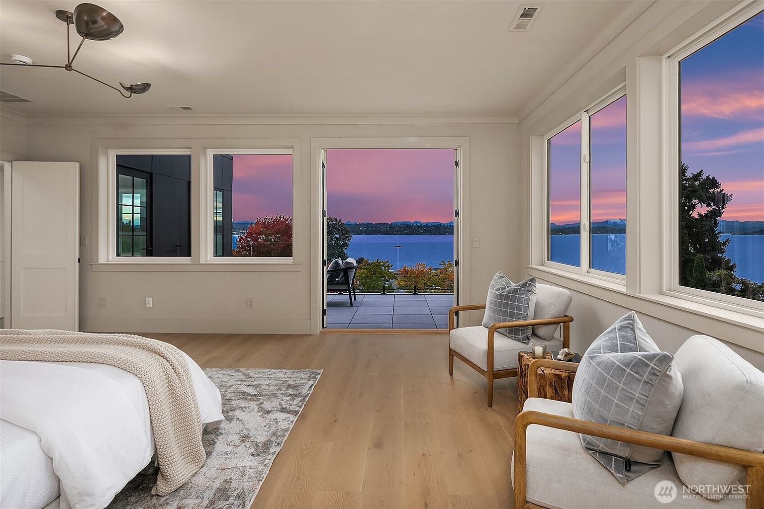 This is a bright and airy primary bedroom with a stunning view of the water and a colorful sunset. The room features hardwood floors, two comfortable armchairs, and large windows that maximize the natural light and scenic views. The decor is minimalist and elegant, creating a serene and inviting atmosphere.