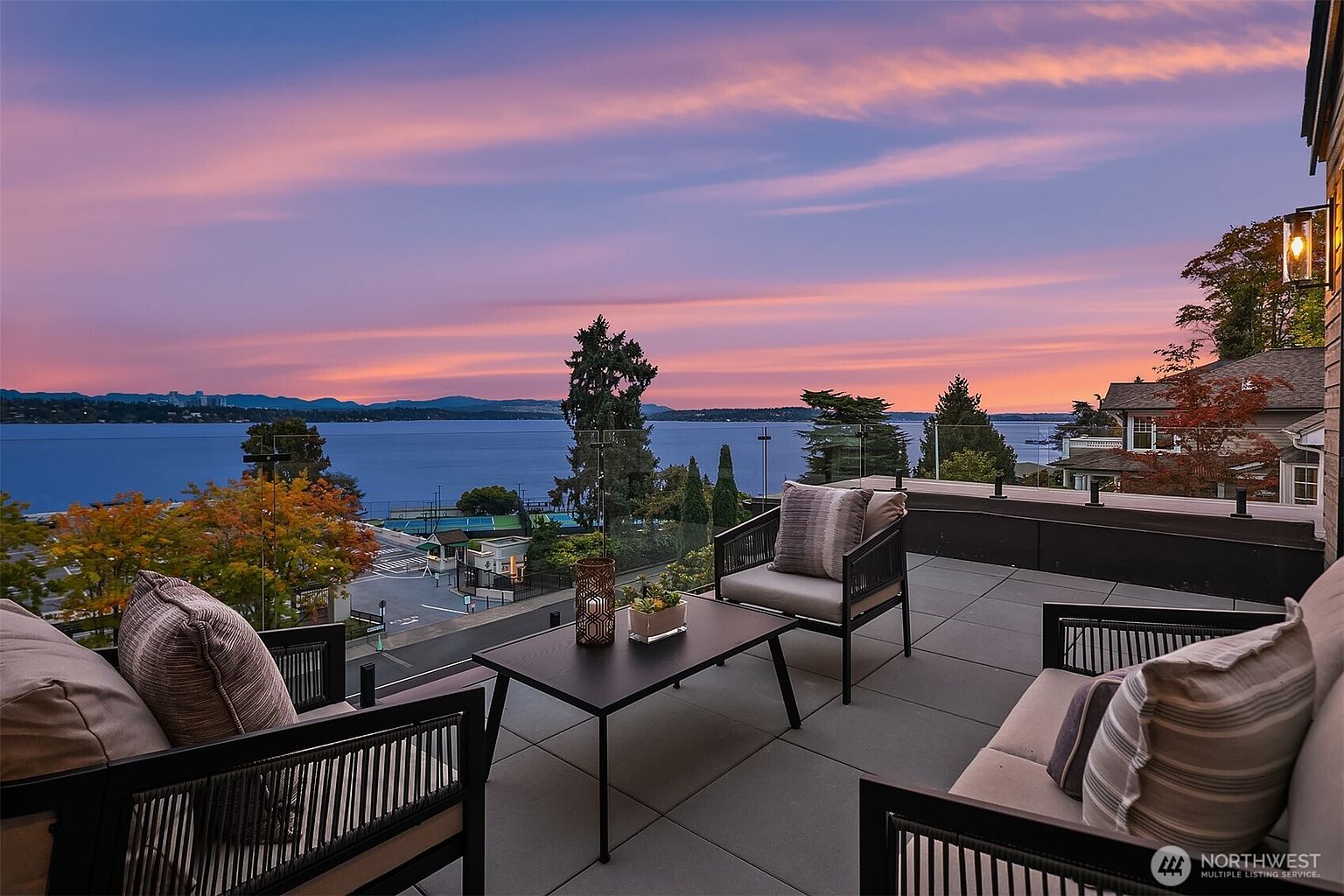 This image showcases a stylish patio or balcony with modern outdoor furniture, including chairs and a table, set against a stunning backdrop of a lake and a vibrant sunset sky. The space is designed for relaxation and entertainment, offering a picturesque view and a sophisticated outdoor living experience. The perspective is from the patio looking out towards the lake.