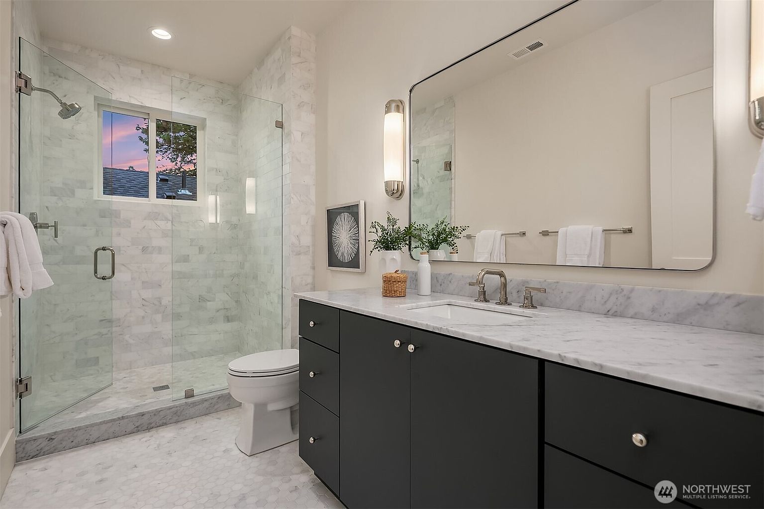This is a well-lit primary bathroom featuring a glass-enclosed shower with marble tiling, a dark gray vanity with a white marble countertop, and a large mirror. The bathroom has a modern and clean aesthetic, with hexagonal floor tiles and sleek fixtures. The window in the shower area provides natural light and a view of the outside.