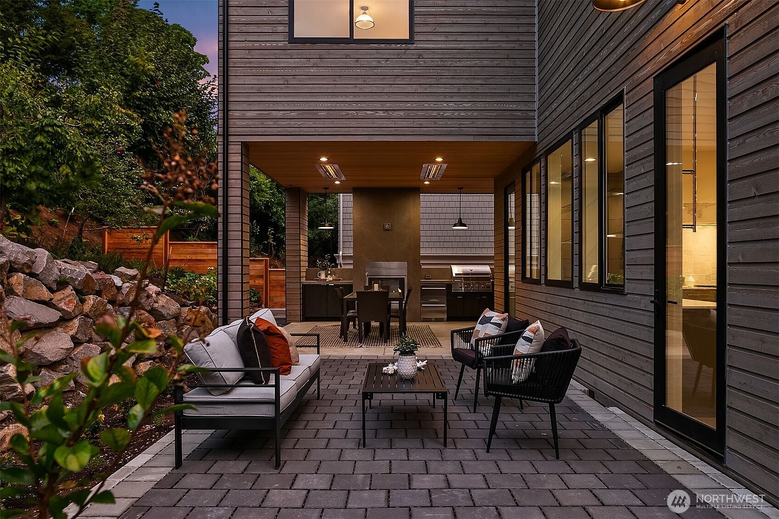 This image showcases a beautifully designed outdoor patio area. The space features comfortable seating arrangements, including a sofa and chairs, arranged around a coffee table on a paved patio. An outdoor kitchen and dining area are visible under a covered section, creating an inviting space for relaxation and entertainment.