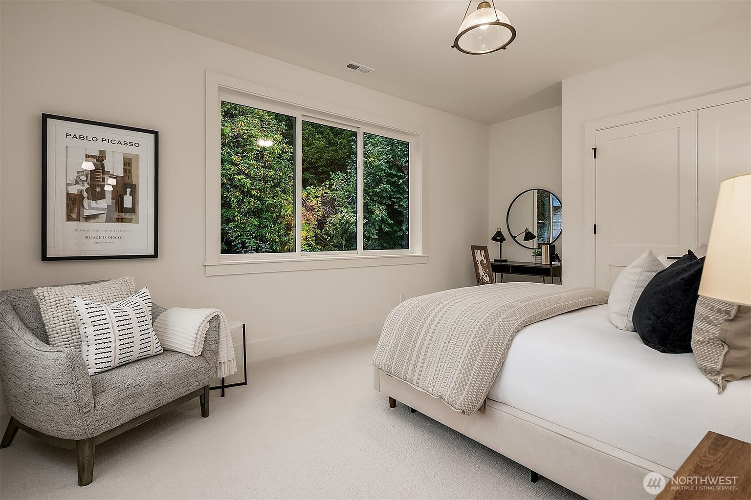 This is a bright and airy bedroom featuring a neutral color palette. A large window provides ample natural light and a view of lush greenery. The room is furnished with a bed, a chair with decorative pillows, and a small desk area, creating a cozy and inviting space.