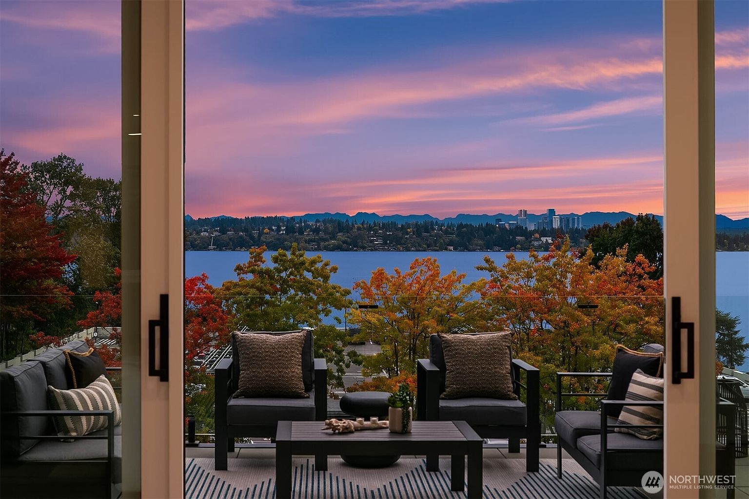 This image showcases a stylish outdoor balcony or patio area, furnished with modern seating and a small coffee table. The view overlooks a serene lake with a colorful sunset sky, framed by trees with autumn foliage. The space is designed for relaxation and enjoying the scenic surroundings, making it an appealing feature for potential buyers.