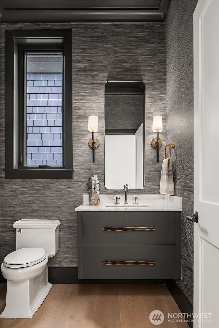 This is a stylish guest bathroom featuring a dark gray vanity with unique drawer pulls and a white marble countertop. The walls are covered in a textured gray wallpaper, complemented by a black-framed window and a matching mirror. The space is illuminated by sconces on either side of the mirror, creating a sophisticated and modern aesthetic.