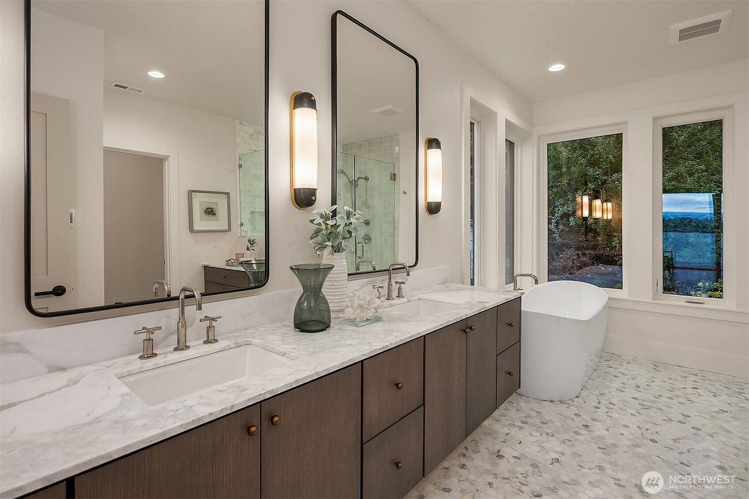 This is a luxurious primary bathroom featuring a double vanity with a marble countertop and dark wood cabinets. Two large mirrors are mounted above the sinks, flanked by modern sconces. A freestanding bathtub sits near a large window offering natural light and a view of the outdoors, complemented by a patterned tile floor.