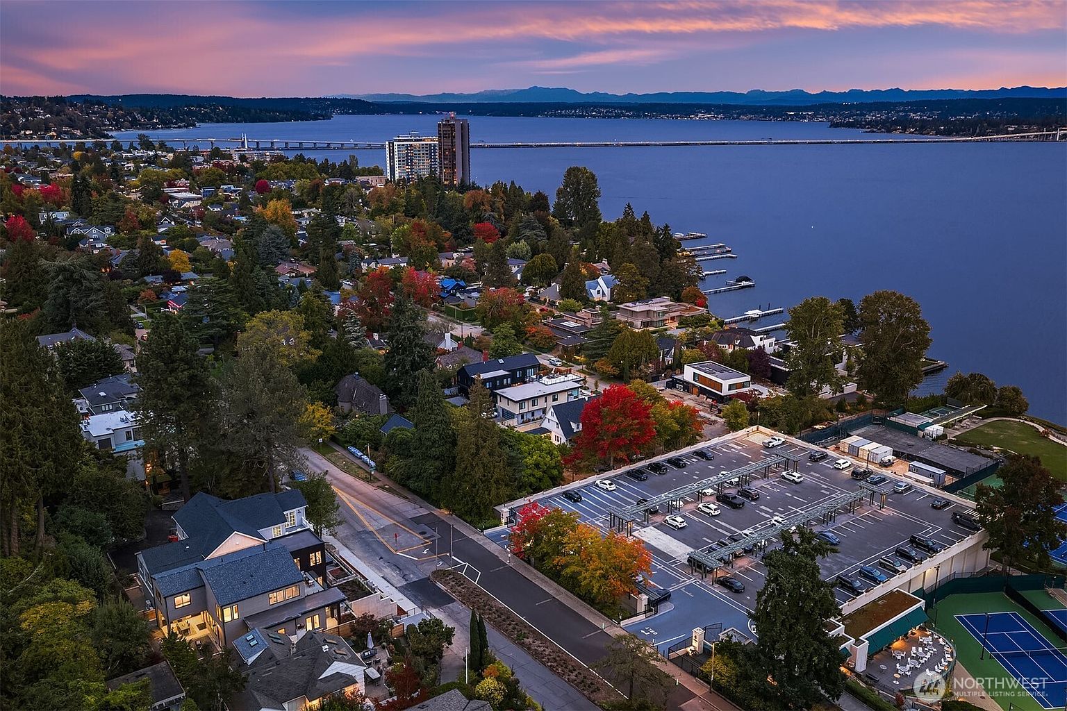 This aerial shot showcases a luxurious waterfront property with stunning views of a lake and distant mountains under a colorful sky. The property features a mix of modern and traditional homes nestled among lush trees with vibrant fall foliage, a parking structure, and tennis courts. A bridge spans the lake, connecting the community to the opposite shore, highlighting the desirable location and upscale neighborhood.